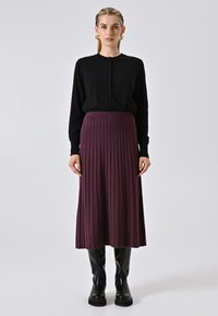 Donna che indossa un cardigan nero con bottoni, una gonna midi ribbed viola, stivali neri fino al ginocchio e orecchini a cerchio dorati, in piedi davanti a uno sfondo neutro.