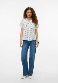 Chemise blanche à manches courtes avec des pois noirs, boutonnée devant, et deux poches poitrine ; associée à un jean bleu évasé et des chaussures blanches.