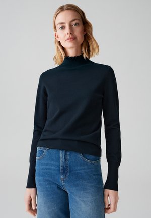 Maglione nero a maniche lunghe con collo alto a costine e un sottile dettaglio arricciato, abbinato a jeans azzurri.