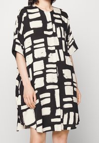 Svart och vit knälång klänning med abstrakt penselmönster, korta ärmar och rund halsringning. Tillverkad av lättviktigt tyg.