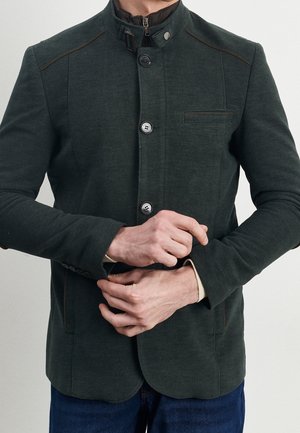 Mann, der den Ärmel einer dunkelgrünen, mit Brusttasche versehenen Jacke mit Knöpfen anpasst, die über einer schwarzen Jacke mit Reißverschluss und blauen Jeans getragen wird.