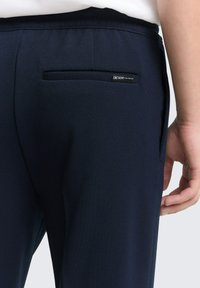 Close-up van een persoon die donkerblauwe broek draagt met een achterste paspelzak en een klein label "Denim Tom Tailor" boven de zak.