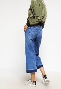 Oliwkowa kurtka bomber z ribbowymi mankietami, w parze z niebieskimi dżinsowymi culottes o średnim stanie z postrzępionymi brzegami oraz srebrnymi trampkami.