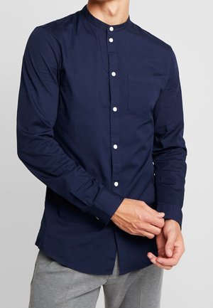 Homme portant une chemise bleu marine à manches longues avec des boutons blancs et une poche poitrine, ajustant le poignet de son bras gauche.