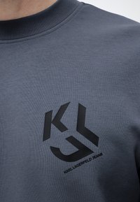 Gros plan d'un sweat-shirt gris avec un logo noir Karl Lagerfeld Jeans comportant les lettres KL et le contour partiel d'un visage.