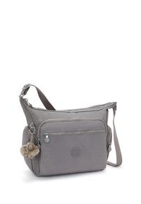 Grau farbene Schultertasche aus Stoff mit mehreren Reißverschlusstaschen, verstellbarem Schultergurt und einem flauschigen Schlüsselanhänger. Rundes Logodesign auf der Vorderseite.