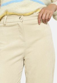 Pantalons en velours côtelé beige avec des nervures verticales, fermeture à bouton et poches latérales. Le tissu a une texture douce et une coupe ajustée.