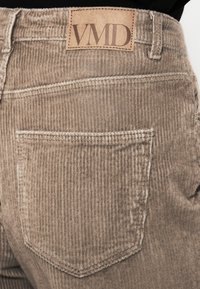 Pantalone od kordura svijetlosmeđe boje s vertikalnom rebrastom teksturom. Na stražnjoj strani nalazi se kvadratna džep i kožni etiket s "VMD" brandom.