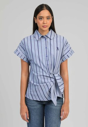 Jeune femme portant une chemise bleue à manches courtes avec des rayures verticales et un nœud noué, associée à un jean bleu, debout devant un fond gris.