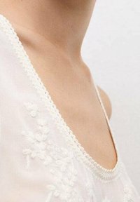 Zbliżenie szyi i ramienia kobiety w białej bluzce z delikatnym kwiatowym haftem i falbanką z koronką.