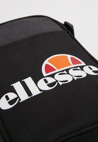 Zaino in tessuto nero con un grande logo bianco "ellesse". Presenta un pannello superiore grigio e un design di mezza-sola arancione e rossa sopra il logo.