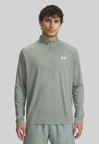 Hellgraues langärmliges Sportshirt mit einem halben Reißverschlusskragen, aus strukturiertem Stoff gefertigt, mit einem kleinen gelben Logo auf der Brust.