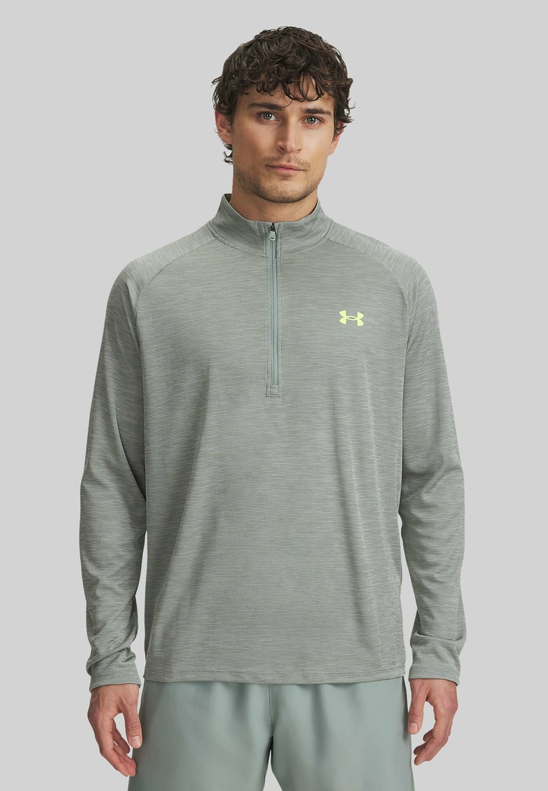 Hellgraues langärmliges Sportshirt mit einem halben Reißverschlusskragen, aus strukturiertem Stoff gefertigt, mit einem kleinen gelben Logo auf der Brust.