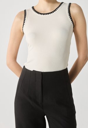 Femme portant un haut blanc côtelé sans manches avec une bordure triangulaire noire et un pantalon noir taille haute sur un fond uni.