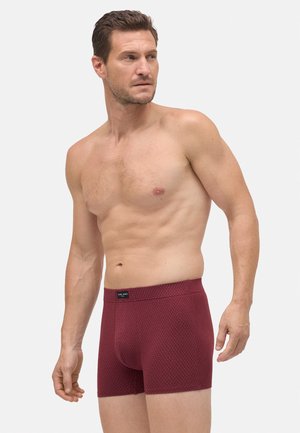 Hombre sin camiseta que lleva ajustados bóxer color burdeos con un diseño de puntos, de pie contra un fondo claro y liso, mirando hacia la izquierda.