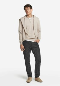 Beżowy sweter z zamkiem przy szyi i luźnym kapturem, zestawiony z ciemnoszarymi dżinsami i jasnobeżowymi butami. Gładka tekstura, swobodny krój, minimalistyczny design.