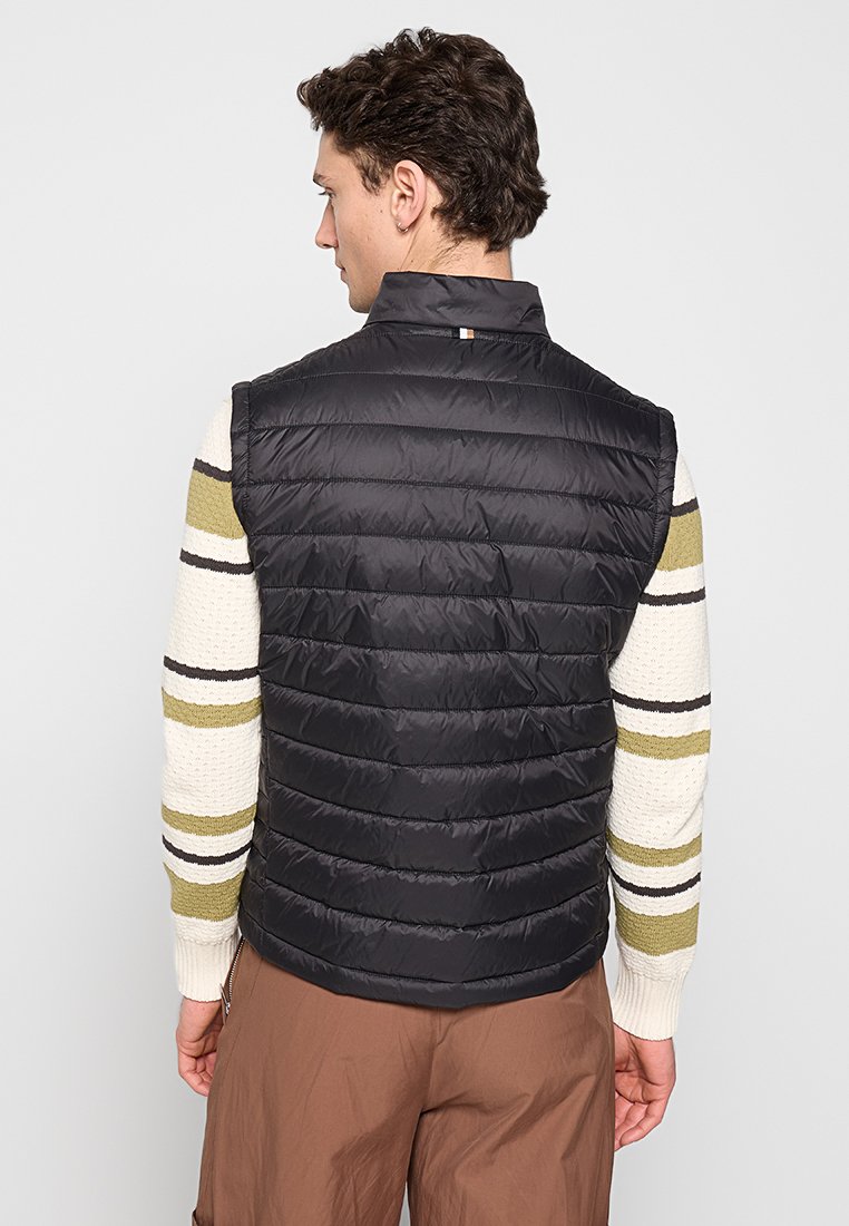 Gilet nero imbottito con trapuntatura orizzontale, colletto alto e un piccolo logo dettagliato sul collo. Indossato sopra un maglione a righe multicolori.