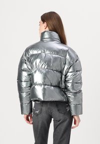 Metallisk sølvfarget pufferjakke med høy krage, med horisontal søm og en cropped design, kombinert med mørkegrå jeans.