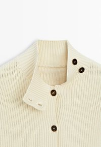 Kremowy sweter dziergany o prążkowanej fakturze, z wysokim kołnierzem i trzema ciemnymi guzikami, dwoma na klapce kołnierza i jednym na korpusie.