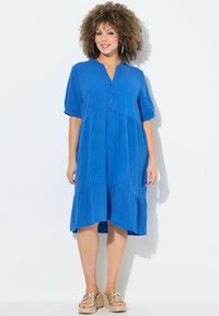 Abito blu a maniche corte con scollatura tipo henley con bottoni, design a strati e tessuto texturizzato. Indossato con sandali con tacco a cuneo e cinturini dorati.