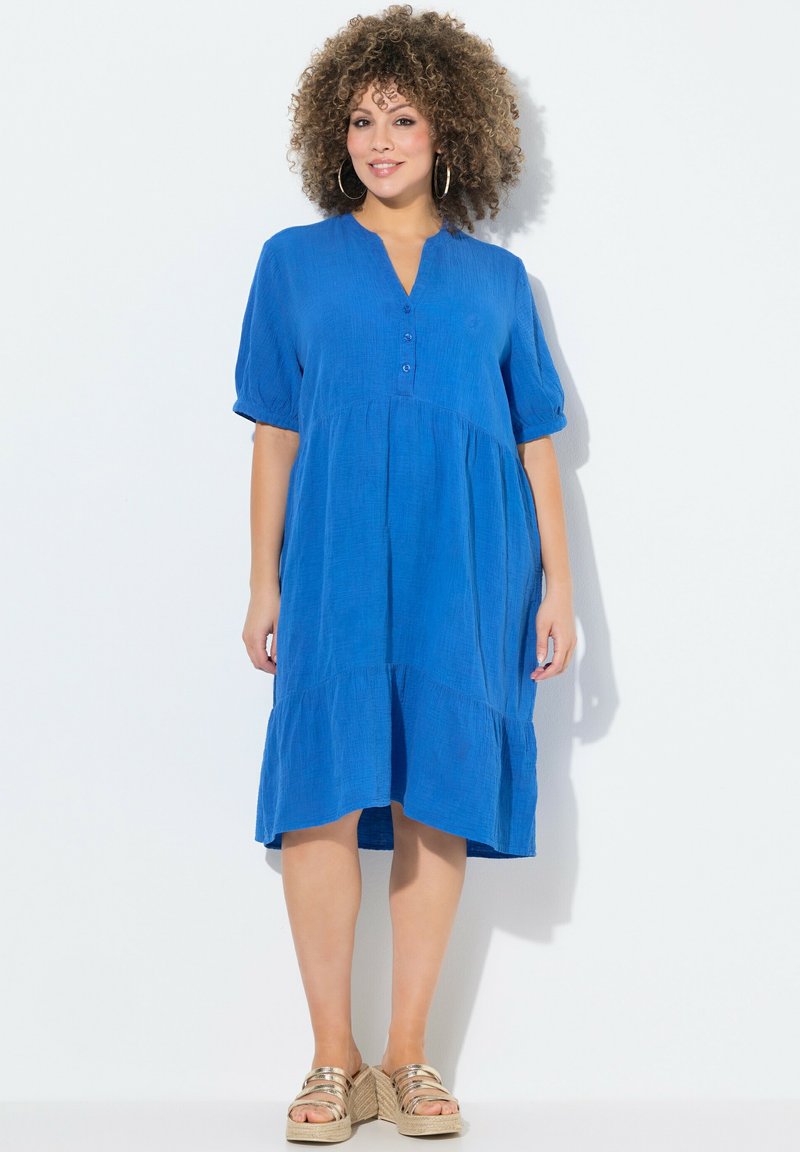 Abito blu a maniche corte con scollatura tipo henley con bottoni, design a strati e tessuto texturizzato. Indossato con sandali con tacco a cuneo e cinturini dorati.