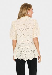 Helles beige Peplum-Bluse mit gewelltem Saum, ausgestattet mit laserperforiertem Lochmuster und kurzen Puffärmeln. Weicher Stoff, feminines Design.