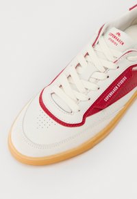 Hvite sneakers med røde detaljer, skinnoverdel, perforert tå og brun gummisåle. Viser "Copenhagen Studios"-logo på siden.