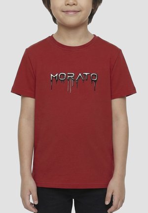 Kind draagt een rood T-shirt met korte mouwen met zwarte en witte druipende tekst "MORATO" op de borst, staat met ontspannen armen.