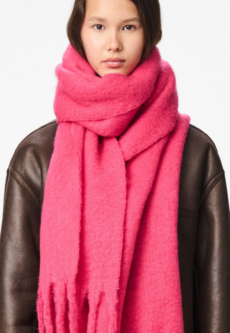 Lyserødt uld tørklæde med blød tekstur og frynser i enderne, viklet om halsen og kombineret med en mørkebrun læderjakke.