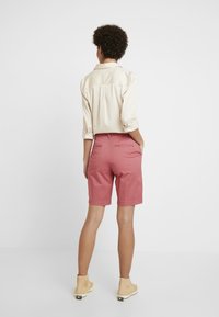 Chemise à manches longues de couleur claire associée à un short rose. Le short présente une texture lisse et des poches arrière, se terminant juste au-dessus du genou.