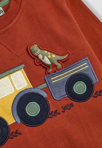 Huppari ruskealla dinosauruspäällyksellä leikkiauton kuvion päällä. Sisältää sinisiä ja keltaisia brodeerauksia pyöreillä renkailla.