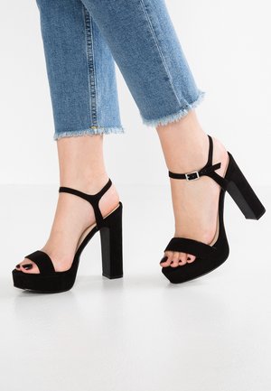 Sorte ruskindssandaler med høje hæle og chunky blokhæl, åben tå og justerbar ankelrem. Har en blød tekstur og en plateau-sål.