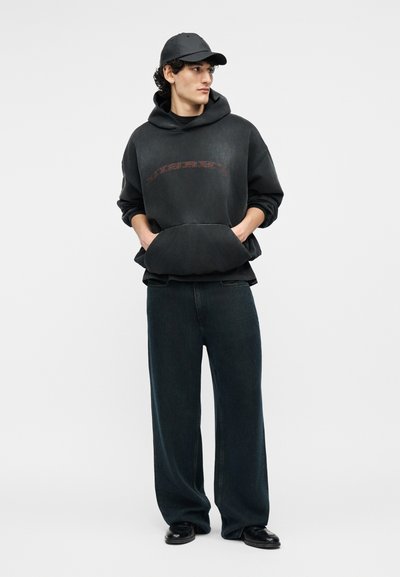 Νέος άνδρας που φοράει μαύρο καπέλο, φαρδιά μαύρη μπλούζα με κουκούλα και ξεθωριασμένη κόκκινη εκτύπωση, φαρδιά σκούρα τζιν και μαύρα παπούτσια, στέκεται με τα χέρια στις τσέπες της μπλούζας με κουκούλα.