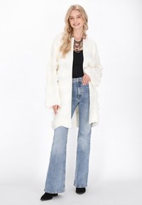 Vit stickad cardigan med texturerade fransar, öppen fram och vida ärmar. Kombinerad med blå utsvängda jeans och svarta ankelboots.