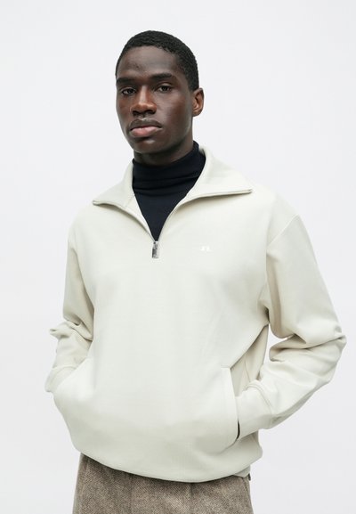 Νεαρός άνδρας με σκούρο δέρμα, φορώντας μπεζ μισό φερμουάρ πάνω από μαύρο πουλόβερ με ψηλό λαιμό και καφέ υφασμάτινο παντελόνι, με τα χέρια στις τσέπες, φόντο ουδέτερου χρώματος.