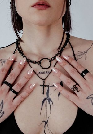 Frau mit schwarzen Ketten und Kreuzanhängern, Tattoo mit der Aufschrift "AMORE" auf der Brust, schwarzen Ringen, nude lackierten Nägeln und schwarzem Trägertop, Hände nahe dem Schlüsselbein.