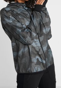 Chaqueta de camuflaje en tonos de gris y azul, con cremallera frontal, capucha y puños elásticos, fabricada con un material ligero y suave.