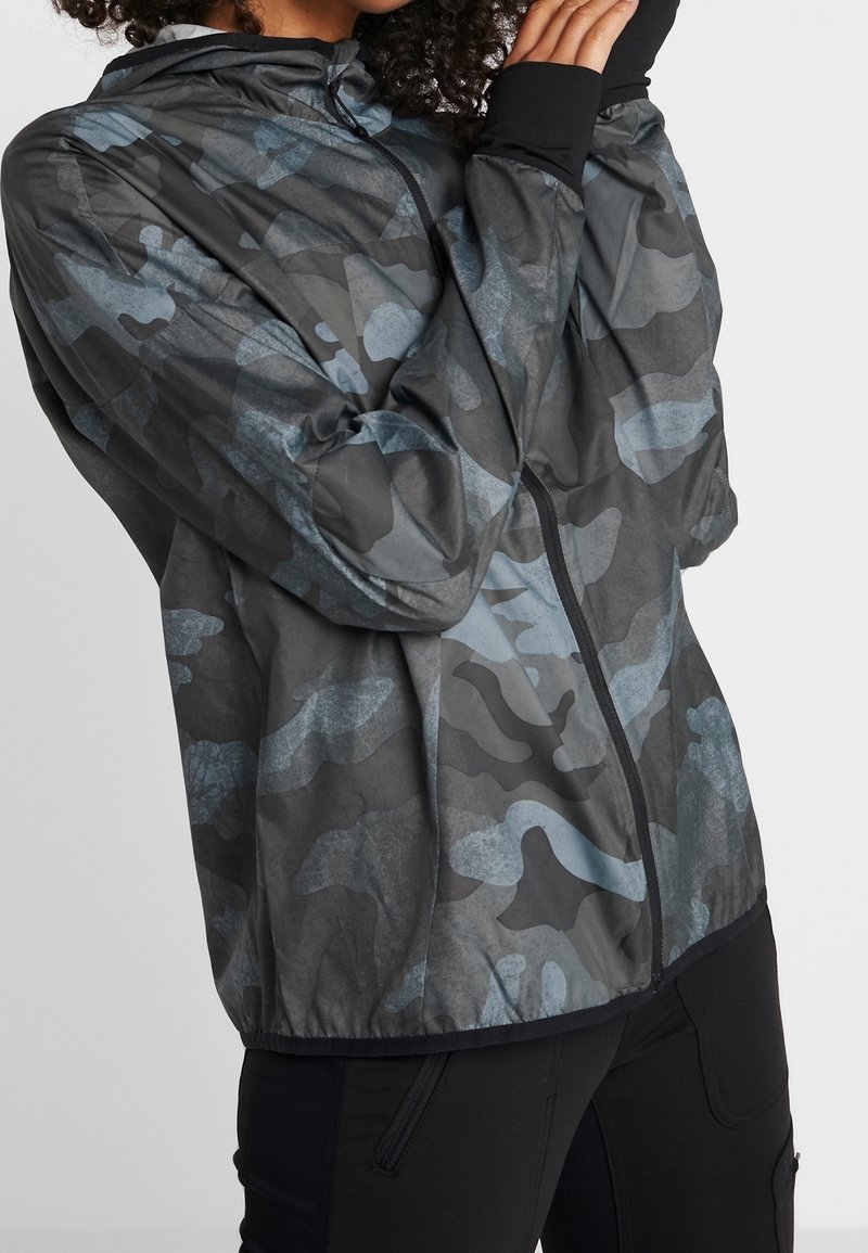 Chaqueta de camuflaje en tonos de gris y azul, con cremallera frontal, capucha y puños elásticos, fabricada con un material ligero y suave.