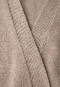 Beżowy kardigan z dekoltem w serek, wykonany z miękkiego, textured dzianiny z prążkowanymi akcentami na brzegach. Zbliżenie pokazuje detale materiału.