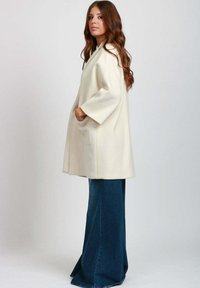 Cappotto beige in misto lana con una vestibilità ampia, maniche larghe e tasche laterali, abbinato a pantaloni in denim blu a gamba svasata per un look contemporaneo.