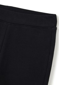 Gros plan sur une ceinture élastique noire et la partie supérieure d'un pantalon noir en tissu, avec des coutures visibles sur un fond blanc.