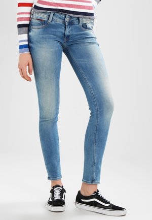 Personne portant un jean slim bleu clair, des baskets noires et blanches, et un pull à manches longues rayé avec des bandes rouges, blanches, bleues et grises.