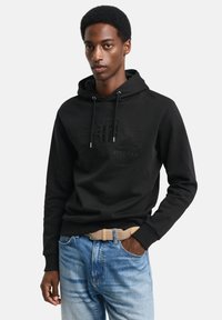 Sweat à capuche noir avec un logo en relief, des cordons de serrage et des poignets côtelés, associé à un jean bleu délavé et une ceinture tissée beige.