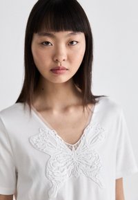 Witte t-shirt met een opvallend geborduurd vlinderontwerp bij de halslijn, met een V-vormige kraag en een losse pasvorm.