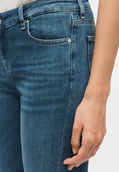 Κοντινό πλάνο του χεριού ενός ατόμου που ακουμπά δίπλα σε μπλε τζιν με ορατές τσέπες και λεπτομέρειες ραφής.