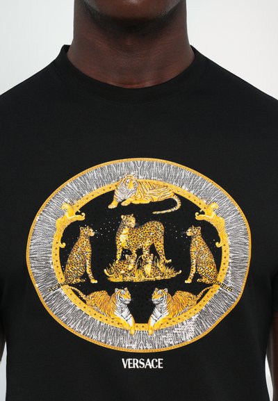 Tricou negru cu un design oval auriu și argintiu care prezintă tigri și leoparzi, înconjurați de modele decorative, cu "VERSACE" brodat dedesubt.