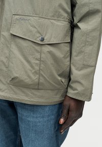 Κοντινό πλάνο ενός χεριού που ακουμπάει στο πλευρό ενός ατόμου που φοράει μπεζ μπουφάν Barbour με εμπρόσθια τσέπη με κουμπιά και μπλε τζιν.