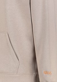 Light beige sweatshirt with a kangaroo pocket, ribbed cuffs, and a visible orange "SBLVL" logo on the sleeve. Soft texture and casual design.