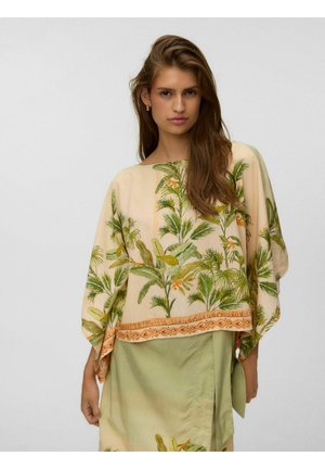 Femme aux longs cheveux portant un haut beige ample avec des motifs de feuilles tropicales vertes et orange et une jupe vert clair sur un fond uni.
