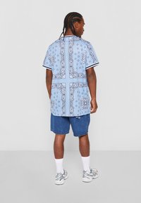Lys blå kortermet skjorte med svarte paisley-mønstre, kontrastfargede striper på kragen og mansjettene, kombinert med denimshorts og joggesko.